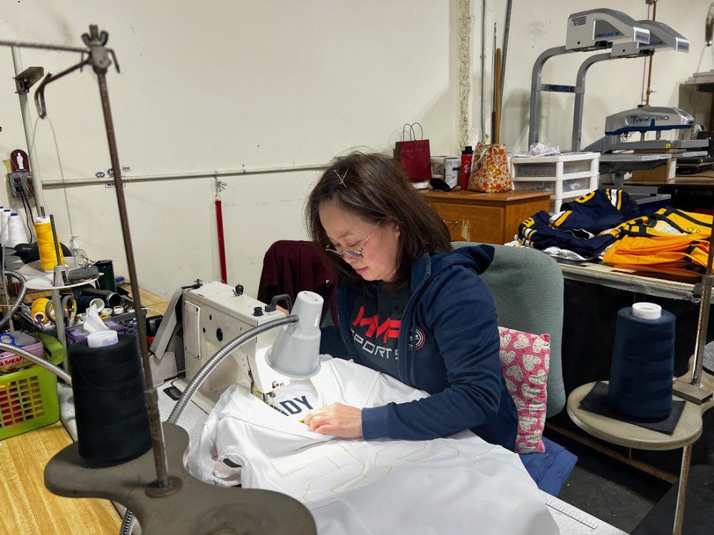 a woman in a blue jacket and glasses operating a sewing machine and sewing a white jersey