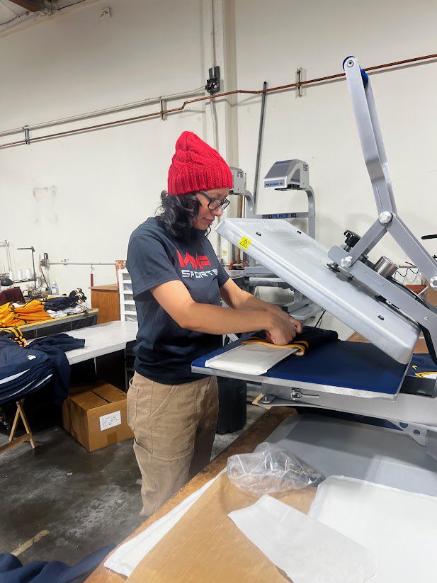 a woman in a red beanie and black MVP Sports shirt operating a heat press machine in a production warehouse