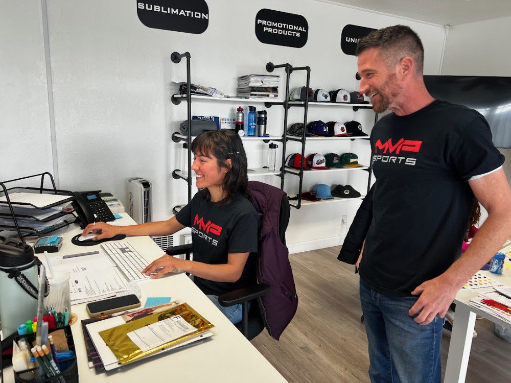 a man and a woman smiling with black MVP Sports shirts working on the computer in an office, with hats and services signage on the wall