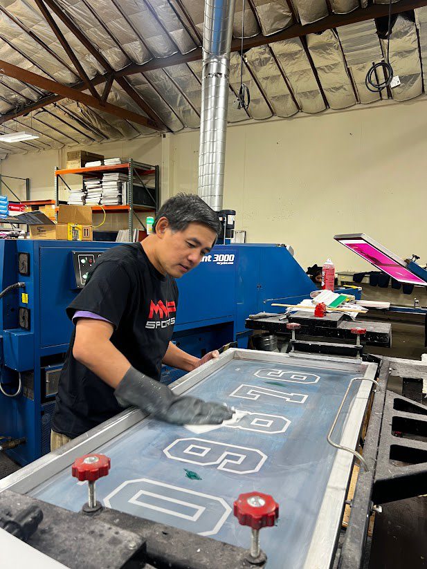a man in a black MVP Sports shirt wiping down a blue and white numbers screen for screen printing
