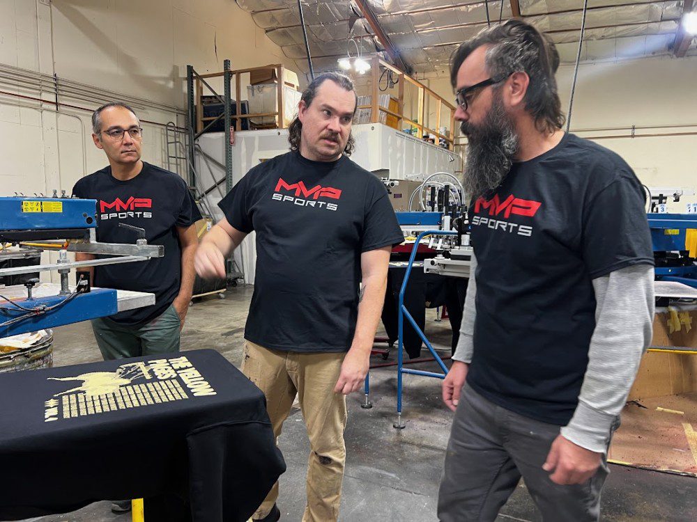 three MVP employees with black MVP Sports shirts talking about a design next to a screen printing machine