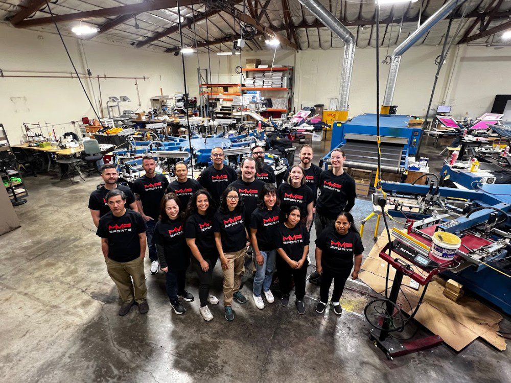 a far away group photo of MVP employees in black MVP Sports t-shirts posing for a photo in the San Leandro MVP production warehouse amongst the screen printing machines