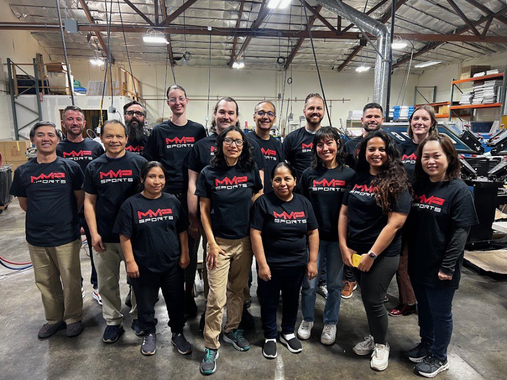 a front-facing group photo of MVP employees in black MVP Sports t-shirts posing for a photo in the San Leandro MVP production warehouse