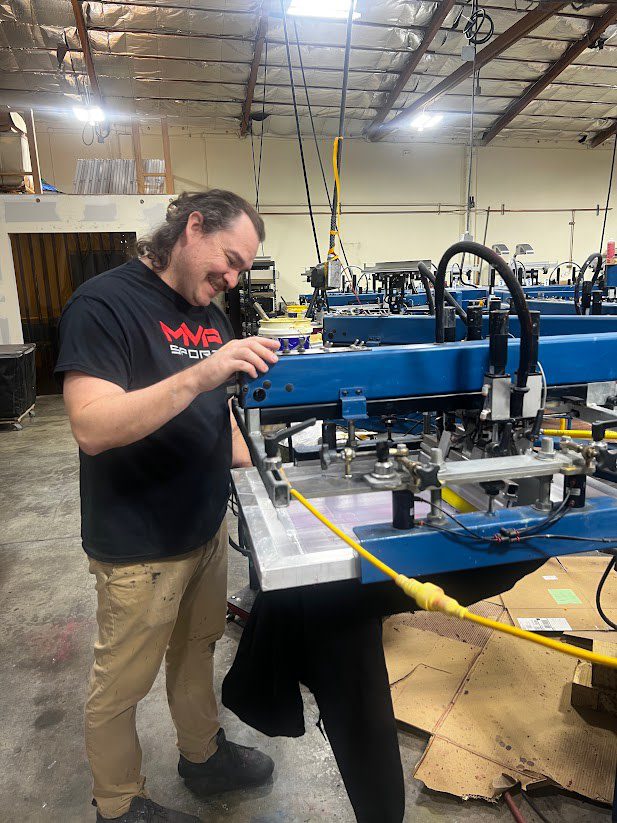 a tall smiling man with a black MVP Sports tee shirt operating a blue screen printing machine in a production warehouse