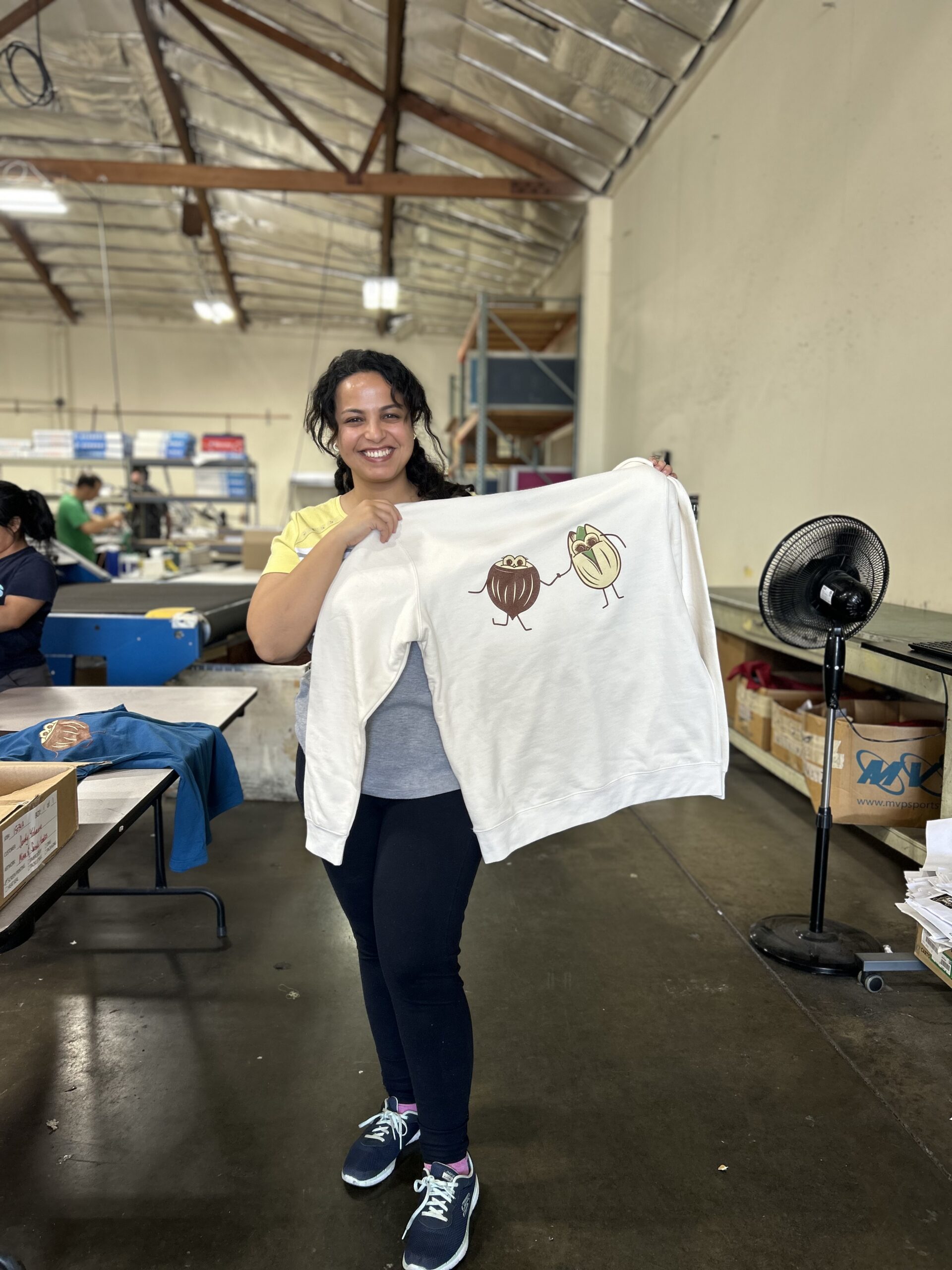 young woman with black hair in a production warehouse holding up a white crewneck sweater with a cute screen printed pistachio and brazil nut design