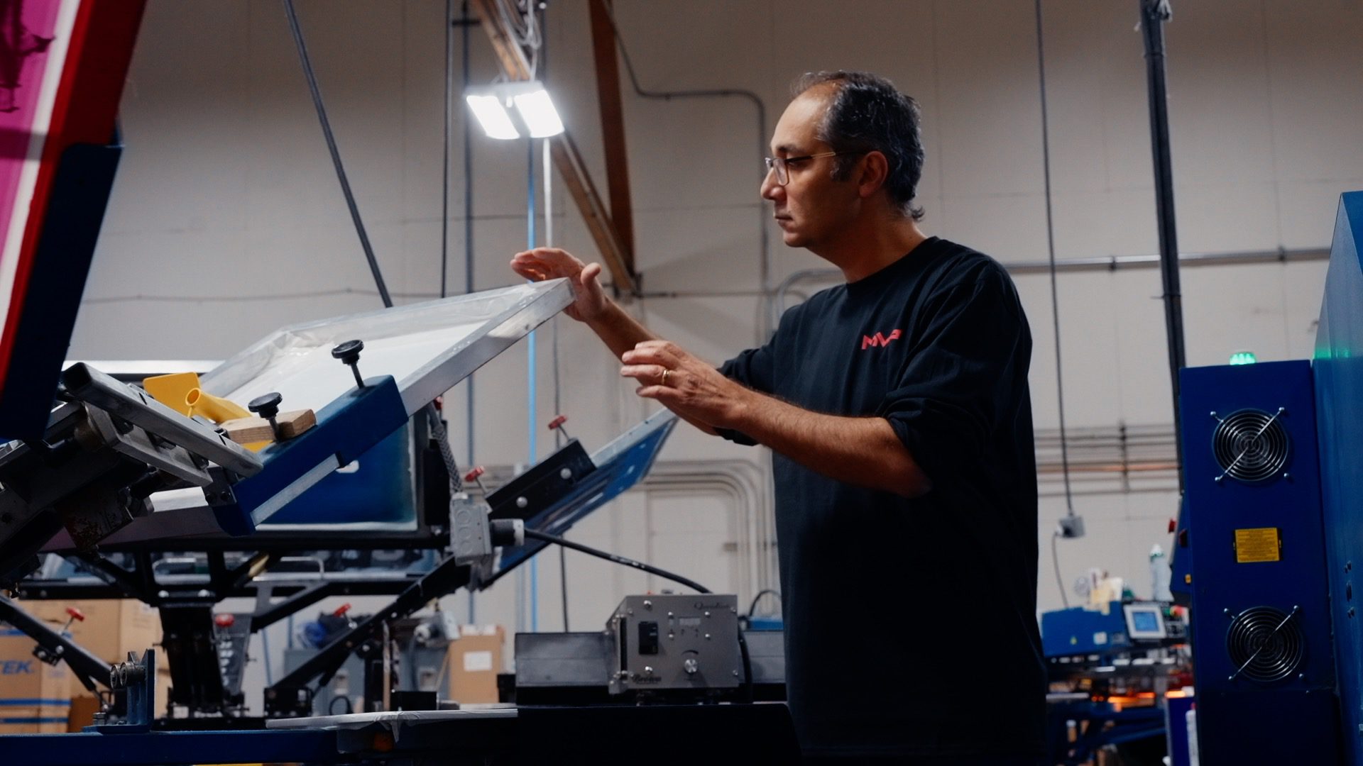 a man with glasses and a black shirt and a red MVP logo operating an automatic screen printing press