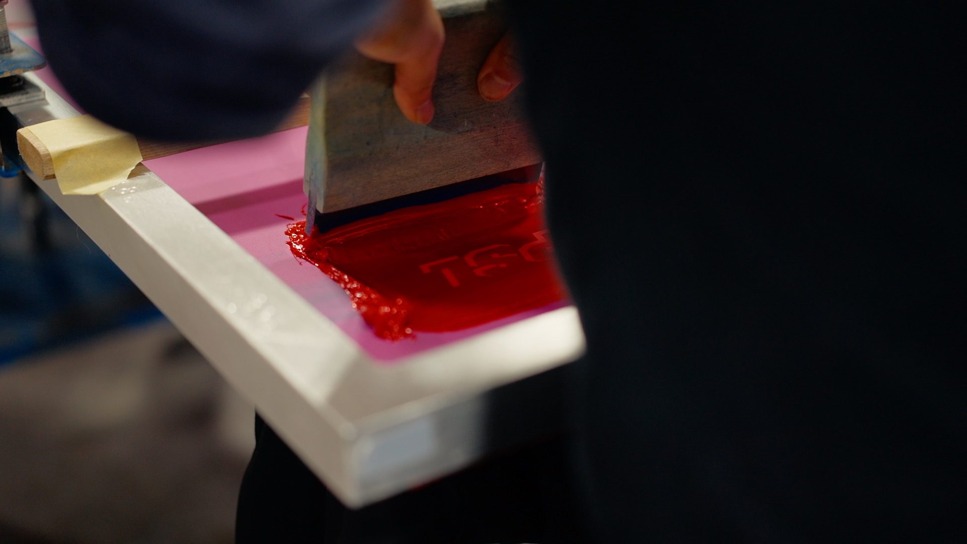 person pressing down a screen printing squeegee, printing a red logo on a black shirt