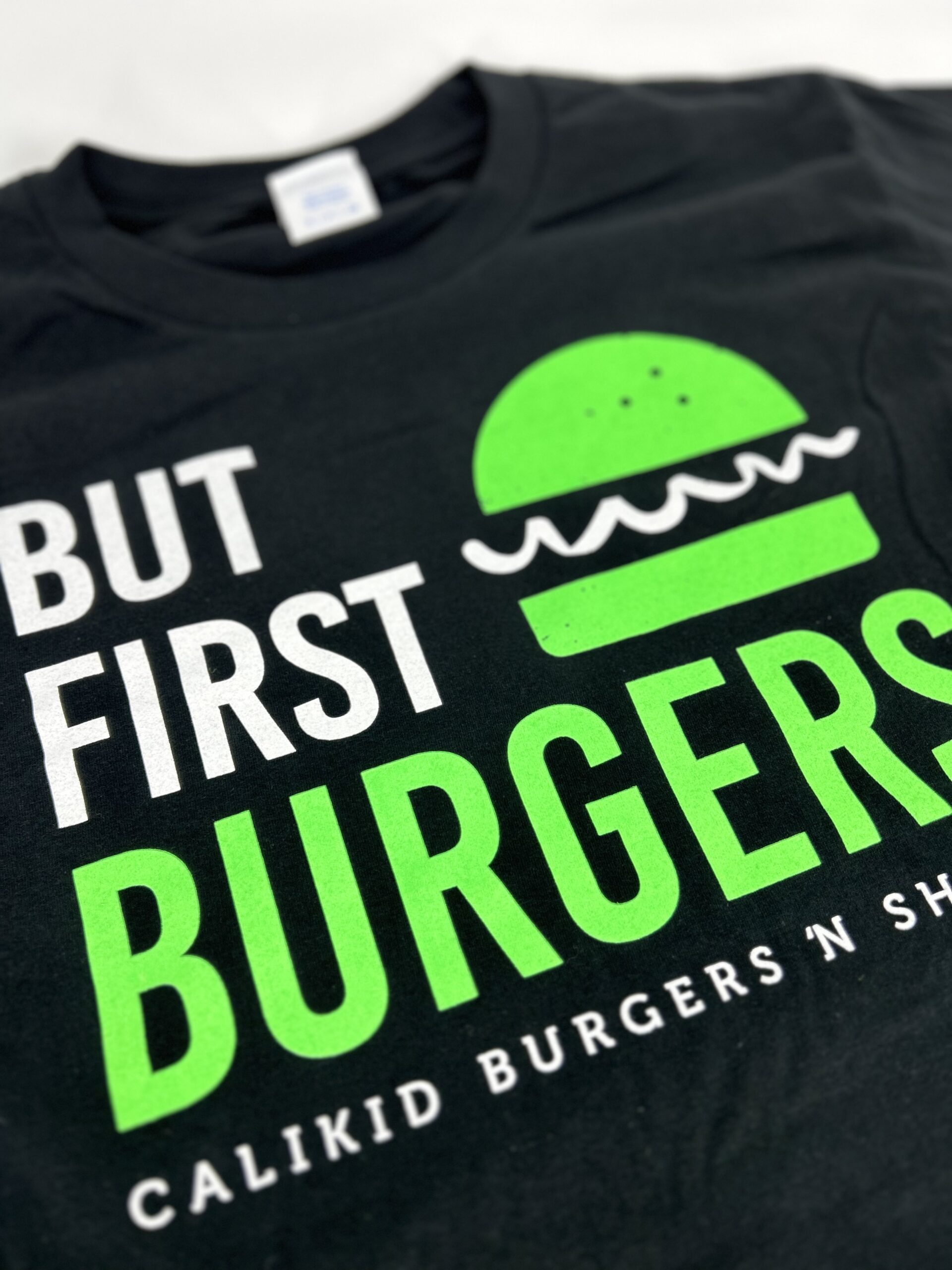 a close up of Cali Kid burgers shirt with green and white design on a black shirt