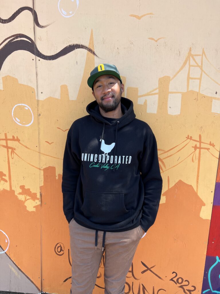 a man in a black Unincorporated Castro Valley hoodie standing against a wall with graffiti