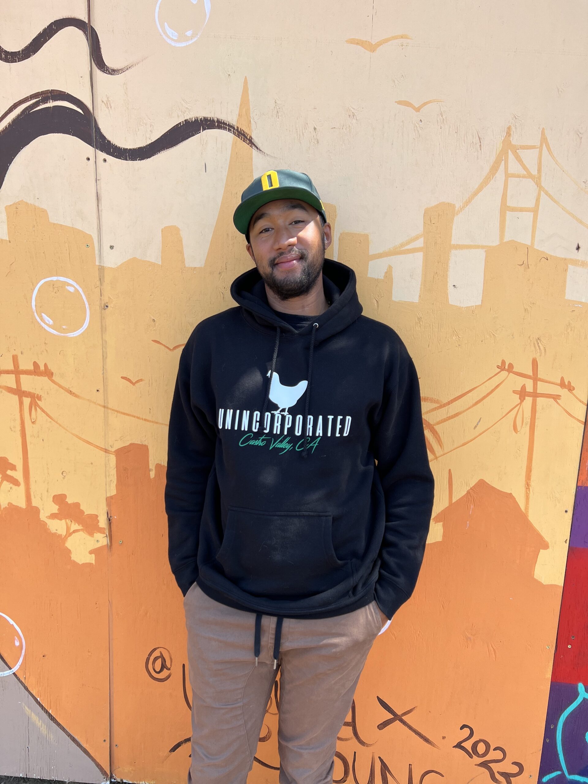 a man in a black Unincorporated Castro Valley hoodie standing against a wall with graffiti
